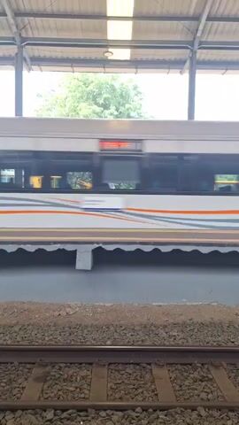 The video shows a train moving quickly past a train platform. The train is white with orange stripes and gray curved lines. The train platform is concrete with a roof supported by metal posts. The ground next to the train track is covered in gravel. Beyond the train platform, there are several pieces of yellow construction equipment. Behind the equipment, there is a tree and sky. The light inside the train station roof is bright.