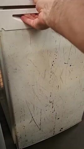 A person opens a refrigerator. The refrigerator is white and appears to be old and scratched. Inside the refrigerator, there are two shelves and a few bottles. One bottle is tall and clear with a red cap, and it is sitting on the top shelf. Another bottle is shorter and darker, and it is on the bottom shelf. A third bottle is visible in the door shelf. The interior of the refrigerator is lit by a warm light.