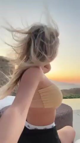 Here is a description of the video:

The video shows a woman sitting outside, facing away from the camera. She is wearing a tan bandeau top and black shorts with a white waistband, and her long blonde hair is being blown by the wind.  The background shows a sunset over mountains, a stone wall, and an infinity pool.