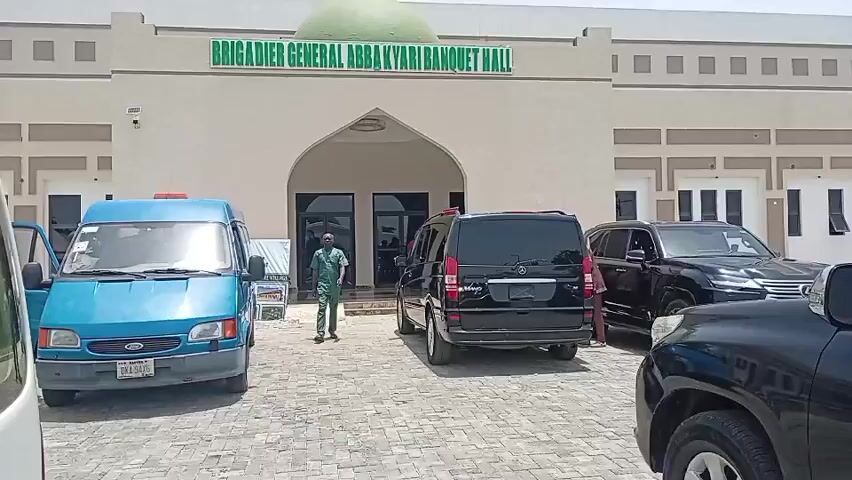 The video captures the exterior of the "BRIGADIER GENERAL ABBA KYARI BANQUET HALL" on a bright, sunny day. The large, tan-colored building features a prominent arched entrance and a green dome on its roof. Several vehicles are parked on a paved area in front of the hall. These include a light blue Ford Transit van with a visible license plate reading "DAA-340HS," a black Mercedes-Benz Viano, and at least two other black SUVs, one of which resembles a Lexus LX.

People are visible in the scene, suggesting arrivals or a gathering. A man dressed in a green uniform walks across the paved area towards the hall's main entrance. Another man, wearing a white tunic, walks towards the black Mercedes. Other individuals are standing near the parked vehicles. The overall scene depicts activity outside the banquet hall, possibly for an event or ceremony, under clear weather conditions.