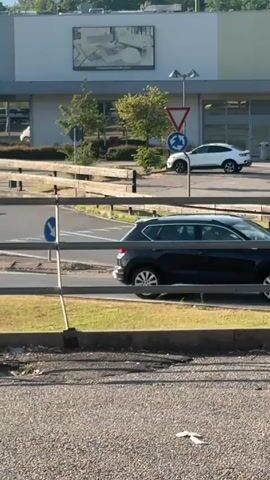 A video shows a street scene with cars driving. In the foreground, there is a gravel surface and a metal railing. Behind the railing, a paved road curves. Several traffic signs are visible, including a yield sign and a roundabout sign. Cars are seen driving on the road, including a white SUV and a small grey car. Trees and a building are in the background. The lighting suggests it is daytime.