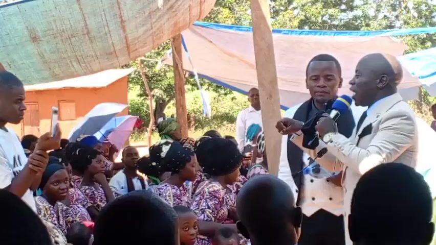 The video depicts a gathering of people, predominantly African, under a makeshift canopy supported by wooden poles. A man in a light-colored suit and tie is speaking into a microphone. Next to him stands another man in a white vest and black tie.  The audience, composed of men, women, and children, is seated facing the speakers. Some women wear matching patterned dresses and headscarves. A person in the crowd is recording the event with a smartphone. A small building is visible in the background. The lighting suggests it's daytime.