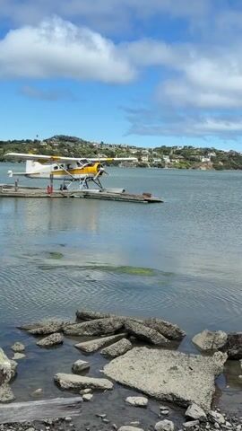 A seaplane sits on a dock on a calm body of water. In the distance, a city skyline is visible along a tree-lined hill. The sky is blue with scattered white clouds. The foreground is rocky and shows the edge of the water.