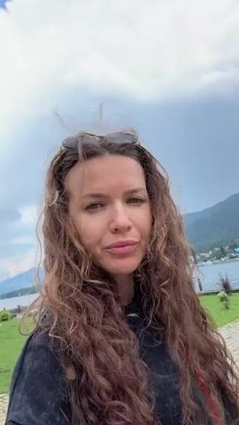 A woman with long, curly brown hair and sunglasses perched on her head is taking a selfie video. She is wearing a dark hoodie with red and green stripes on the strap of a bag visible over her shoulder. The background shows a cloudy sky, a body of water with mountains in the distance, and a grassy area with lounge chairs and a paved walkway. The woman turns her head to the right, revealing more of the landscape behind her. The wind is blowing her hair.