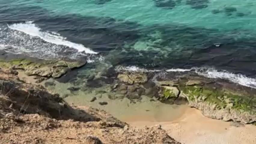 The video shows a view from atop a cliff looking down at a small cove. The cove has a sandy beach surrounded by rocky outcroppings with green moss. The ocean water is turquoise in some areas and dark blue/black in others. Small waves are breaking on the rocks and beach. The camera slowly zooms in towards the ocean. There are no people or animals visible. The environment appears to be a coastal area.