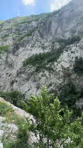 The video shows a mountainous landscape under a partly cloudy sky. The focus is on a deep canyon with steep, rocky cliffs covered in patches of green vegetation.  At the bottom of the canyon, dense green foliage is visible. A narrow road winds along one side of the canyon. In the later part of the video, a person is seen walking on the road.