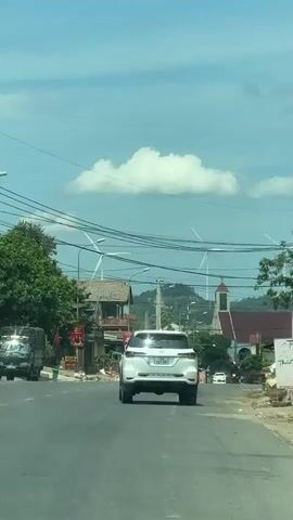 This video shows a view from a vehicle traveling down a road in Tân Lập Commune, Vietnam. The road appears to be a main thoroughfare in a semi-rural area, with buildings and trees lining the sides.

In the foreground, a white SUV is driving away from the camera. Behind it, another white vehicle, possibly another SUV or a pickup truck, is also visible. Further down the road, a military-style green truck is traveling in the same direction. Several other vehicles and motorbikes are scattered along the road.

The scene is set under a bright, partly cloudy sky, suggesting it is daytime, likely late morning or early afternoon. The sun is casting shadows, indicating a clear and sunny period within the day. The presence of wind turbines on a distant hill provides a notable detail about the landscape and the region's energy infrastructure. A building resembling a church with a red roof and a steeple is also visible among the other structures.

Power lines crisscross the sky, a common sight in many parts of Vietnam. The buildings along the road range from simple structures with wooden elements to more modern-looking establishments. A red flag with yellow text is displayed on one of the buildings. The overall atmosphere appears to be one of everyday activity on a road with a mix of residential and commercial elements, set against a backdrop of natural scenery and renewable energy sources.

The license plates of the vehicles are partially visible but are not clear enough to read specific details. One visible license plate on the white SUV appears to start with "72A". The truck in front has a license plate that might start with "93C". The weather is fair and warm.