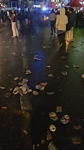 A person stands on a street at night littered with trash. Several other people are walking around on the street. A woman in a white jacket and black pants smiles at the camera. There are broken bottles, cans, and other trash on the ground. There are buildings and streetlights visible in the background. A police car with flashing lights can be seen in the distance.
