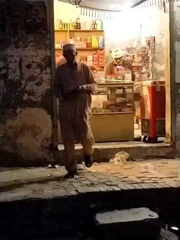 The video depicts a night scene outside a small, brightly lit shop in what appears to be a South Asian setting, likely Dera Chahal, Pakistan.

In the foreground, an elderly man wearing a light-colored shalwar kameez and a white prayer cap walks from left to right, passing by the shop entrance. The ground outside the shop is a rough, uneven brick surface, with some litter visible. The exterior walls of the surrounding buildings are made of brick and appear worn.

The shop itself is well-lit from within, contrasting with the dark exterior. Inside, a glass display counter holds various items. Behind the counter, a man with a beard, wearing a dark shirt, appears to be the shopkeeper. To his right, another man wearing a light-colored cap is also present, looking at the items in the shop. Shelves packed with bottles and packaged goods line the back wall of the shop, and a red upright freezer or cooler is visible to the right of the entrance.

The activity suggests a typical evening or nighttime street scene, with people moving past and a shop operating. The artificial lighting inside the shop clearly indicates it is dark outside. No specific legible text is visible other than what might be a brand logo on the red cooler.