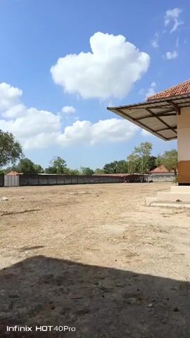 The video shows an outdoor scene on a sunny day with a clear blue sky and scattered white clouds. In the foreground, there is a dry, dusty ground. In the middle ground, there is a covered area with a red roof supported by poles, under which a group of people are seated, facing the camera, alongside several parked motorcycles. Behind this shelter, a long, brick wall runs across the frame, and beyond that, there are trees and buildings. To the right of the shelter, a single black motorcycle is parked. A white structure is visible in the lower right corner of the frame. A watermark in the bottom left corner indicates "Infinix HOT 40 Pro". The camera is stationary.