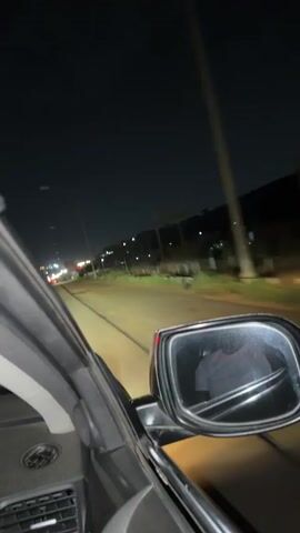 The video shows a view from inside a car driving at night. The camera is positioned near the driver's side mirror, capturing the passing roadside scenery in the mirror and through the side window. The night sky is visible above. The roadside includes trees, foliage, buildings, and utility poles lit by streetlights. Other car headlights are also visible. The interior of the car is dark. The video is somewhat blurry due to the motion of the car.