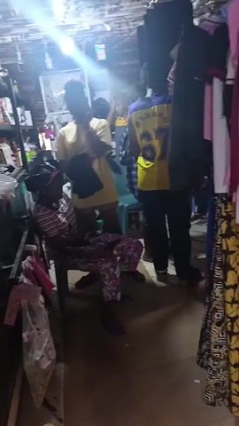 The video shows the interior of what appears to be a small clothing store or market stall. A woman is seated on a plastic chair, looking at her phone. She's wearing a striped top and floral pants. Several people are standing around, including a person in a yellow jersey with the number "67" on the back. There are clothes hanging on racks to the right of the frame. Shelves stacked with various items are visible to the left. The lighting in the space is somewhat dim, with bright overhead lights creating a glare in the camera.