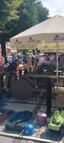 This video depicts an outdoor market scene. Several people are present, browsing items displayed on tables and on the ground. In the foreground, there are boxes containing various goods, a brown suitcase, and what appears to be a propane tank. Behind these items, there is a metal railing and some stacked tables and chairs. The background shows more stalls, trees, and buildings, suggesting a busy urban environment. A large white umbrella with logos is prominently featured overhead. Some individuals are interacting with the merchandise, while others are conversing. The overall atmosphere appears casual and lively, possibly a flea market or street fair.