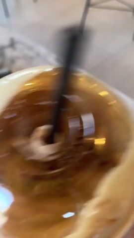 A person's hand holds a clear plastic cup filled with a frothy, dark brown beverage and a black straw. The cup is covered with a clear plastic lid that has a slight dome shape. Foam from the drink spills over the edge of the lid. The background is blurred and appears to be an indoor setting with tables and chairs. The video is shaky and the camera movement suggests the person holding the cup is walking or moving.