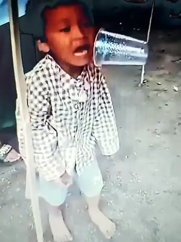 The video shows a young child wearing a checkered shirt and shorts. The child's mouth is open wide and their head is tilted back. A clear plastic cup is positioned on the child's cheek, near their ear. The background appears to be outdoors, with other people visible in the distance. The ground is dirt or gravel. The child's skin appears flushed, possibly from exertion or heat. The overall scene suggests a moment of playful activity or performance.