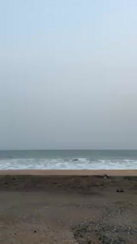 The video shows a wide shot of a beach and ocean. The sand is a darker color and appears slightly damp. Waves are crashing on the shore. The sky is pale and overcast. There are no people or other distinct objects visible.