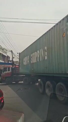 A green shipping container with "CHINA SHIPPING" written on its side is being transported on a truck on a street. A motorcycle is visible on the left side of the frame. The scene appears to be outdoors with power lines visible overhead. The video is short and shows the truck from the side as it moves.