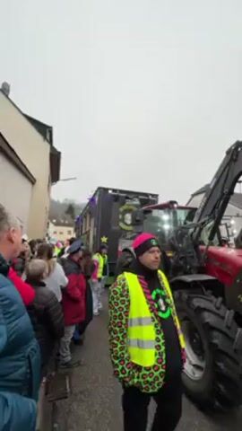 The video captures a scene of a parade, possibly a carnival or festival, taking place on a street lined with buildings. A crowd of people, including adults and children, stand on the sidewalk watching. A large black trailer decorated with lights and a green logo is being pulled by a red tractor down the street. A person wearing a neon yellow vest with a pink animal print stands near the tractor, facing forward. Some members of the crowd are dressed in costumes. The weather appears overcast, with a gray sky.