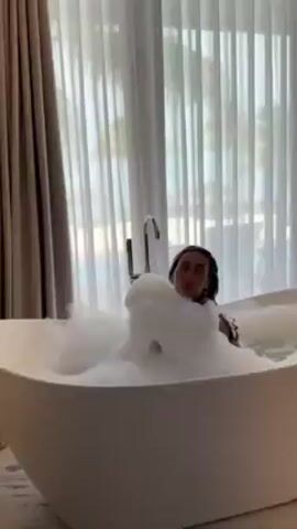 A young woman relaxes in a luxurious white bathtub filled with thick, white bubbles. She is facing towards the window, which is covered by sheer white curtains and reveals a beach scene outside. The room is well-lit, and a modern chrome faucet is visible on the edge of the tub. The woman’s dark hair is wet and slicked back. She calmly enjoys her bath, seemingly unwinding.