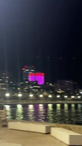 A night time video shows a blurred cityscape across a body of water. The city is illuminated with various lights, including some that appear to be pink, blue, and yellow. There are buildings and what looks like a waterfront promenade. In the foreground, there is a white railing and two white benches. The water reflects the city lights. The video is shaky and out of focus.