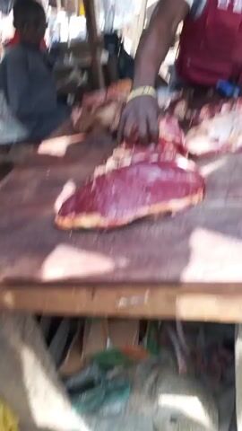 The video shows a man, likely a butcher, working at a stall in what appears to be an outdoor market. He is standing behind a wooden table covered with pieces of raw meat, using a large knife to cut them. The man is wearing a white t-shirt, a red vest with writing on it, and a baseball cap. Above him, a faded umbrella provides shade. There are other vendors and customers visible in the background. Underneath the table, there is some discarded trash and debris.
