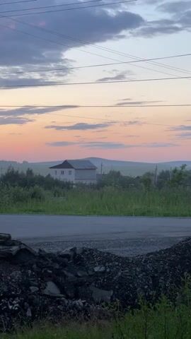 The video pans across a rural landscape at dusk. In the foreground, a pile of dark, rough debris sits near a paved road. Beyond the road, lush green vegetation and trees are visible. In the middle ground, a white house with a dark roof is situated amongst trees and foliage. Further in the distance, rolling hills are shrouded in a light mist. The sky above is a gradient of soft pinks, oranges, and purples, with scattered clouds and visible power lines stretching across the frame. The overall impression is one of a quiet, serene countryside evening.