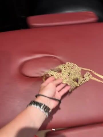 The video shows a person's hand holding a gold lace mask in the foreground. In the background, there is a red massage table and a dark shelf filled with books and decorative items. The shelf contains several books, a black box with a blue item inside, a decorative golden apple, a black box filled with red roses, an hourglass filled with golden sand, and a black ornate mask under a glass dome. The lighting in the room appears to be dim, with some reflections visible on the shelf surfaces.