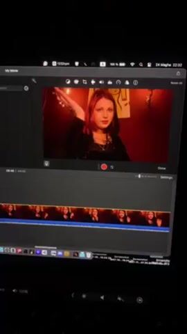 A dark-haired person with red and blonde dyed hair is in the frame of a video editing software. The person is in a red lit room, wearing a black outfit, with chains around their neck. There are multiple cuts of the same clip edited into a timeline below the frame.