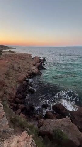 A vertical video shows a rocky coastline with the ocean on the right and the sky on the left. The sun is setting, casting warm colors across the horizon. The water is a deep blue, with some areas of turquoise near the shore. Waves crash against the rocks, creating white foam. There are no people or man-made objects visible in the video.