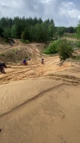 The video shows a group of people riding dirt bikes in a sandy, hilly area with trees in the background. The terrain is uneven and appears to be a motocross track or a similar off-road riding area. Several riders are visible on their bikes, navigating the sandy slopes and tracks. The sky is cloudy. The camera perspective is dynamic, moving with the riders and capturing the action from various angles. Some riders are going uphill, while others are descending. There are tire tracks visible in the sand, indicating recent activity. The overall scene depicts an active and potentially challenging off-road dirt biking environment.