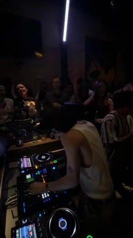 Here's a description of the video:

The video shows a DJ performing in a crowded, dimly lit bar or club. He is standing behind a DJ console, manipulating the equipment with his hands. The crowd is dancing and raising their hands, enjoying the music. There are several people visible in the background, including some standing near the DJ booth and others on the dance floor. The lighting is dynamic, with spotlight effects emphasizing the DJ and occasionally illuminating parts of the crowd. The overall atmosphere is lively and energetic.