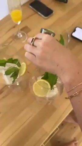 A hand wearing a gold ring and bracelet pours liquid from a measuring cup into a glass. The glass contains ice, a lemon slice, and mint leaves. There are two similar glasses next to it on the table, each containing ice, mint, and a lemon slice. There is a glass of orange juice and two cell phones on the table. A small yellow package of lemon drops is also visible. The table is made of wood.