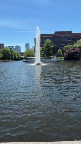 A high fountain sprays water in the middle of a pond. A white boat is visible behind the fountain. In the background, there are buildings and trees. The sky is blue with some clouds. The video pans to the left, showing a gazebo by the water and more buildings. The camera then pans back to the right, returning to the fountain.