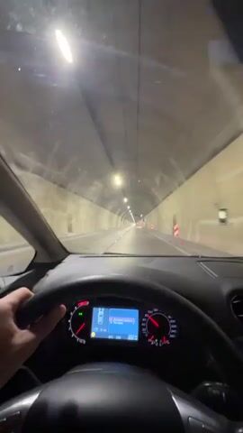 The video provides a first-person perspective from inside a car driving through a well-lit tunnel. A hand is visible holding the left side of the steering wheel. The car's dashboard is prominent, featuring a speedometer, tachometer, and a central digital display. The speedometer indicates a speed of approximately 80 km/h, and the tachometer needle hovers around 2500 RPM. The digital screen shows the external temperature as "20.0 C", the time as "13:24", and other readings including "51 km" and "20593 km".

The tunnel, located in Đurmani, Montenegro, has light-colored, curved walls and a ceiling illuminated by numerous bright lights. The road ahead appears straight and clear, with distant vehicle lights visible. Along the right side of the tunnel, emergency exits or service areas are marked, and some traffic cones are present, indicating possible road work or a designated lane.