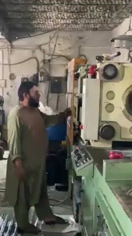 A man wearing a long green shirt and sandals operates an industrial injection molding machine in what appears to be a factory setting. He presses a button on the machine, and it opens. The man pulls out a small, green, plastic piece. After removing the piece, he presses a button again and the machine closes. The factory has a corrugated metal ceiling, and wires and electrical boxes can be seen on the walls. The floor appears to be dirty or wet. Other machinery and equipment are visible in the background.