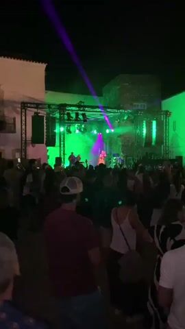 A vertical video captures a lively outdoor nighttime event, possibly a concert or festival. A band performs on a stage illuminated by vibrant green and purple lights, with spotlights creating beams that cut through the darkness. A crowd of people, visible from behind, stands in front of the stage, appearing to enjoy the performance. Some individuals are holding up their phones, likely recording or taking pictures. The environment suggests a public gathering, with buildings visible in the background, their white facades illuminated by the stage lighting. The overall atmosphere is energetic and celebratory.