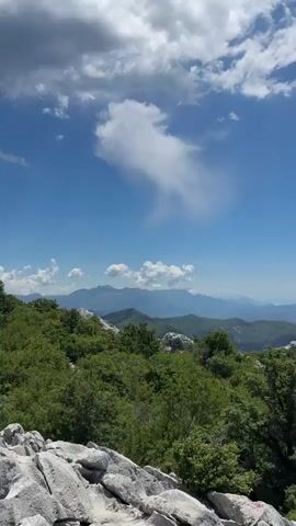The media file captures a breathtaking panoramic view of a mountainous landscape under a bright, partly cloudy sky. In the foreground, large, weathered grey rocks dominate the scene, their rough textures clearly visible. Beyond the rocks, lush green trees and dense foliage spread across the slopes, creating a vibrant contrast.

Further in the distance, a series of rolling mountains fade into a hazy blue, suggesting a vast and expansive terrain. The sky is a striking cerulean, punctuated by dramatic white clouds, one of which has a wispy, elongated shape that draws the eye. The overall impression is one of natural beauty and serenity.

The time of day appears to be daytime, likely mid-morning or early afternoon, given the bright sunlight illuminating the landscape and casting distinct shadows. The weather seems pleasant and clear, ideal for enjoying the vista. The location cues, such as the rugged rocky terrain and the distant coastal mountains, point towards a Mediterranean or Adriatic region. No people or objects are visible, emphasizing the raw, untamed nature of the scene. The image evokes a sense of awe and peacefulness, inviting the viewer to contemplate the grandeur of the natural world. No text is visible in the media file.