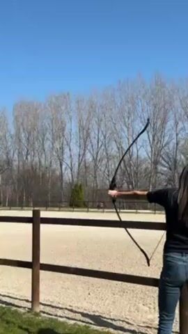 A woman is drawing a bow and arrow in an outdoor setting. The woman is facing away from the camera, and she is wearing a black shirt and blue jeans. She is standing in front of a wooden fence, and behind the fence is a large, sandy area, likely an equestrian arena.  There are leafless trees in the background under a clear blue sky.