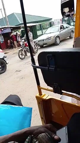 The video shows a view from inside a yellow auto-rickshaw, looking outwards. A person in a white shirt with some text on the back is sitting in the auto-rickshaw. In the foreground, a black seat is visible. Outside the auto-rickshaw, on the left side, a silver sedan is parked at an angle, facing the left. A man in a white robe and a cap is riding a red motorcycle, also facing left, near the silver car. Further to the left, there are buildings and other people. In the background, to the right, other vehicles are visible, including a white truck with people in the back. The sky is overcast. The auto-rickshaw appears to be stationary or moving slowly.