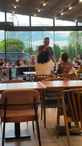 A man stands and speaks into a microphone in front of a group of people seated at tables in a restaurant. He is wearing a patterned short-sleeved shirt and white pants. The restaurant appears to be enclosed with large windows and has an outdoor view. There are lights strung across the ceiling. The people at the tables are diverse in age and gender. They are looking at the speaker and some appear to be engaged in conversation. The tables are wooden and have chairs around them. Some of the tables have plates and glasses on them. The overall atmosphere seems casual and social.