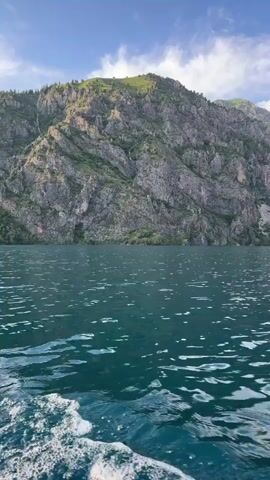 The video shows a body of water with ripples, likely from a boat. In the background, there are tall, rocky mountains with some greenery on their slopes. The sky is blue with scattered clouds. The camera appears to be moving, looking towards the mountains and across the water. There are no people or discernible objects on the water other than the ripples. The overall scene is a natural landscape.