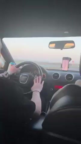 The video shows the interior of an Audi passenger car being driven on a highway. The driver is a person wearing a black t-shirt and a watch. Their hands are on the steering wheel. A phone with a GPS navigation app is mounted on the dashboard. The video then shifts focus to the view out the window, showing a flat, grassy landscape under a clear, pale blue and pink sky. The camera is moving, indicating the car is in motion. A white vehicle is seen passing in the distance. A gray guardrail runs alongside the highway.