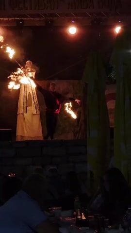 Two performers are on a stage in front of an audience at night. One performer, dressed in a long, white, medieval-style gown, stands on stilts and juggles flaming objects. The other performer, wearing dark clothing with red accents, juggles flaming objects at a lower level. The stage is lit by spotlights and has a backdrop that appears to be decorated with natural elements. The overall atmosphere is one of a nighttime performance or festival, likely outdoors given the presence of umbrellas in the background. The text at the top of the image appears to be in Cyrillic, possibly indicating the name of the event or festival. The audience is seated in the foreground, and the lighting suggests it is dark outside, consistent with a nighttime event. The flames create a dramatic and energetic visual effect.