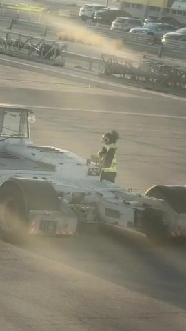 A white airport tow tractor drives on an asphalt surface, kicking up dust. A person wearing a neon yellow vest and dark hair pulled back is visible in the driver's seat. The tractor has the words "Quality Airport Services" printed on its side. In the background, there are several parked cars, metal barriers, and airport infrastructure. The sun appears to be low in the sky, casting a warm light and creating lens flare.