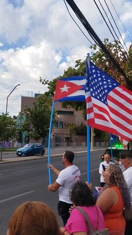 A group of people hold an American and a Cuban flag as they stand near a road with cars passing by. The sky is cloudy and there are trees in the background. A building with glass windows is also visible in the background. One man is holding the American flag on a blue pole. Another man is holding the Cuban flag on a blue pole. Some of the people are taking pictures with their phones. One of the men is wearing a white shirt that says "Queremos #Libertad." There are power lines in the upper left corner of the frame.