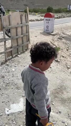 A young child stands on the side of a road, holding a toy construction vehicle. In the background, several motorcycles are parked. A milestone indicates 12 kilometers to Maan. The environment is mountainous with a cloudy sky. The video pans to show the motorcycles, the mountainous landscape, and the road.