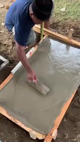 A man in a blue shirt and a black Muslim cap is kneeling outside, smoothing wet cement within a rectangular wooden frame. He is using a trowel to even out the surface. A level is visible beside him, and other tools are nearby. The area around the frame is dirt. The man is wearing flip-flops.
