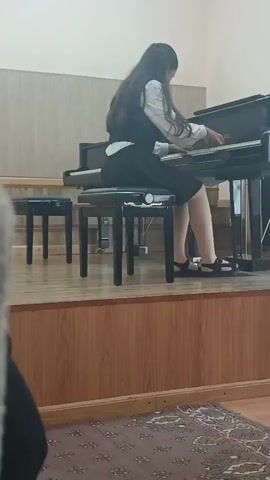 A young girl with long dark hair, wearing a white shirt and a black vest and skirt, sits at a black grand piano on a wooden stage. She is playing the piano, her hands on the keys. To her left are two small black stools. The stage has a wooden front with horizontal slats. In front of the stage is a patterned rug. The background is a light-colored wall with some darker paneling. The lighting appears to be artificial. The video is short and shows the girl playing the piano.