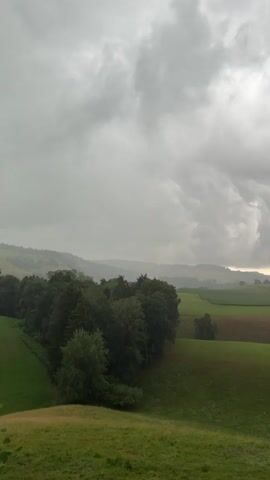 The video shows a rural landscape under a cloudy sky. The scene is dominated by rolling green hills, with patches of trees scattered throughout. In the distance, a small village with buildings and a church steeple is visible. The overall atmosphere is serene and natural, with a hint of approaching rain suggested by the heavy clouds. There are no people or animals visible in the video. The video pans slowly from left to right, revealing more of the landscape and the village.