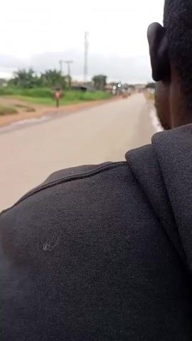 A person wearing a black hooded sweatshirt is facing away from the camera. The camera is looking over their shoulder from a slightly low angle. They appear to be on the back of a moving vehicle, likely a motorcycle or scooter. The road is paved and appears to be in a rural or semi-rural area. In the distance, there are some buildings, trees, and a tall communication tower. Several other vehicles are visible on the road, including a yellow auto-rickshaw and a motorcycle. The sky is overcast.